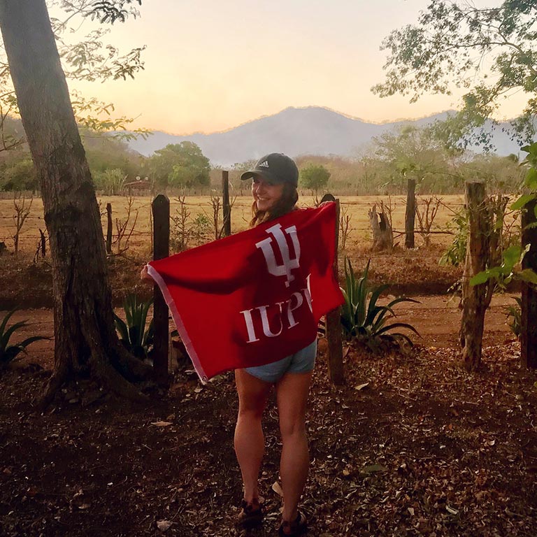 A student in the countryside holds an IUPUI flag.